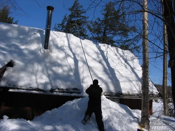 Человек убирает снег с крыши дома специальным скребком с длинной ручкой