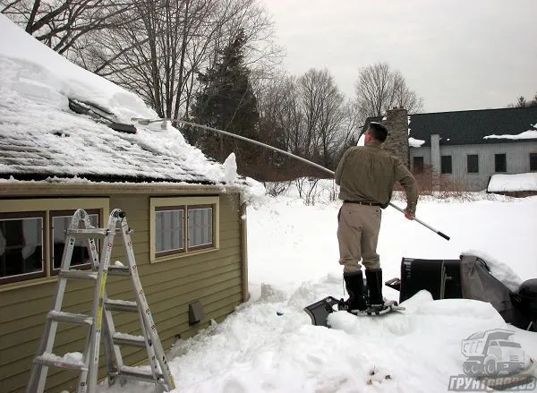 Человек убирает снег с крыши дома специальным скребком с длинной ручкой, стоя на возвышенности