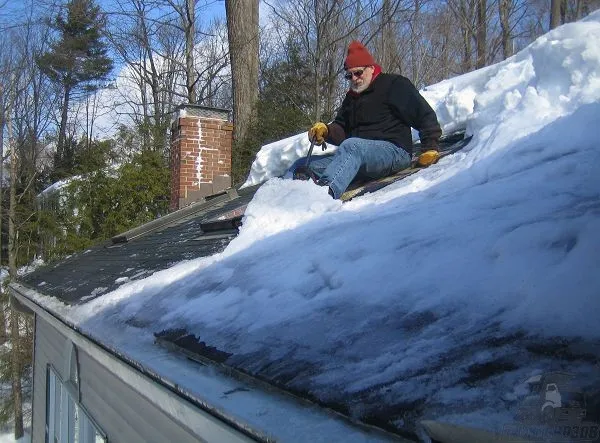 Человек убирает снег с крыши дома, сидя на крыше