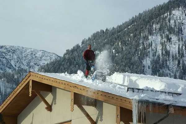 Человек убирает снег с крыши дома, будучи на крыше, при помощи снегоочистителя
