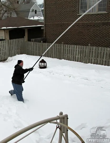 Человек убирает снег с крыши своего дома специальным скребком с длинной ручкой