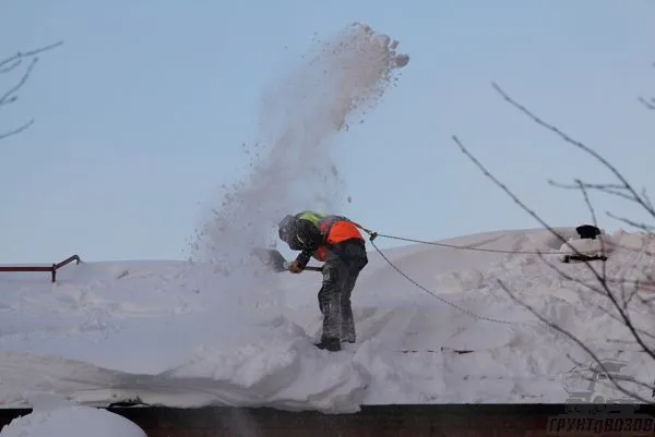 Человек убирает снег с крыши дома, стоя на крыше